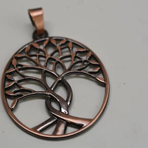 May include: A copper-colored pendant featuring a tree of life design. The pendant has an oval shape with a detailed tree silhouette inside. The tree's branches and leaves are intricately cut out, and the pendant has a loop for a chain.