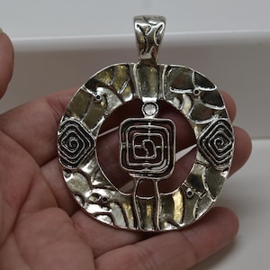May include: Silver-toned pendant with an abstract design. The pendant features a circular frame with a textured surface and a central square element. Two spiral designs are on either side of the square.