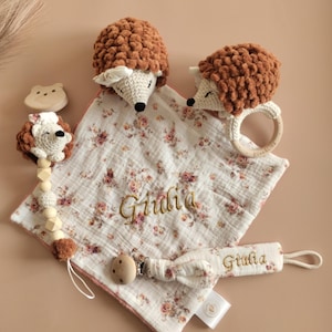 May include: A baby gift set with a personalized muslin blanket, a wooden teether, a crocheted hedgehog rattle, and a crocheted hedgehog with a wooden bead necklace. The blanket is white with a floral pattern and the name "Giulia" embroidered on it.