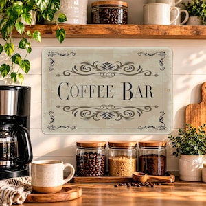 May include: A rectangular metal sign with a beige background and dark gray decorative scrollwork borders. The text COFFEE BAR is printed in a dark gray serif font in the center. The sign features rounded corners and small mounting holes.