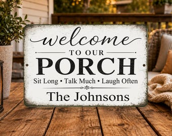 Könnte beinhalten: Ein rechteckiges, verwittertes weißes Schild mit schwarzem Text: „welcome TO OUR PORCH Sit Long • Talk Much • Laugh Often The Johnsons“. Das Schild steht auf einem Holztisch, mit einer Veranda und Grün im Hintergrund.