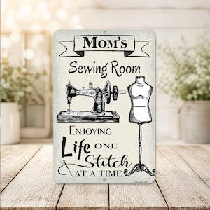 May include: A rectangular metal sign with the words "Mom's Sewing Room" above a sewing machine and dress form illustration. The sign also includes the phrase "Enjoying Life One Stitch At A Time."