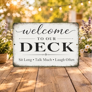 May include: A rectangular sign with the words "welcome TO OUR DECK" in black and white. Below the text are the words "Sit Long • Talk Much • Laugh Often". The sign has a distressed, vintage look.