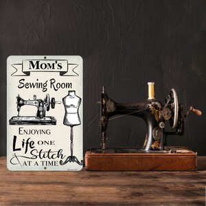 May include: A vintage sewing machine with gold accents sits on a wooden surface next to a metal sign. The sign reads "Mom's Sewing Room" with illustrations of a sewing machine and dress form, and the text "Enjoying Life One Stitch At A Time."
