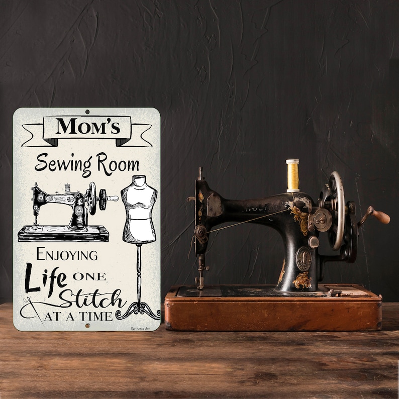 May include: A vintage sewing machine with gold accents sits on a wooden surface next to a metal sign. The sign reads "Mom's Sewing Room" with illustrations of a sewing machine and dress form, and the text "Enjoying Life One Stitch At A Time."