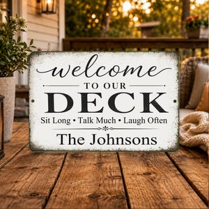 May include: Rectangular, distressed-style sign with the words "welcome TO OUR DECK" in black and white. Below, it reads "Sit Long • Talk Much • Laugh Often" and "The Johnsons". The sign is on a wooden surface.