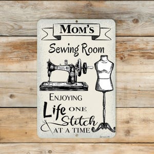 May include: A rectangular metal sign with the words "Mom's Sewing Room" above a sewing machine and dress form illustration. The sign also includes the text "Enjoying Life One Stitch at a Time."
