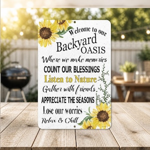 May include: A rectangular metal sign with the words "Welcome to our Backyard Oasis" and other phrases like "Listen to Nature" and "Relax & Chill." The sign is decorated with sunflowers and green leaves. The sign is on a wooden table.