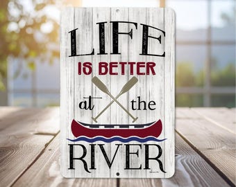 Könnte beinhalten: Rechteckiges Schild mit einem Hintergrund in Holzoptik. Der Schriftzug "LIFE IS BETTER at the RIVER" ist in Schwarz und Burgunderrot gehalten, mit gekreuzten Rudern und einer roten Kanu-Grafik.
