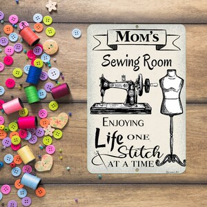 May include: A metal sign with the text "Mom's Sewing Room" and the phrase "Enjoying Life One Stitch At A Time." The sign features illustrations of a vintage sewing machine and a dress form. Colorful spools of thread and buttons surround the sign.