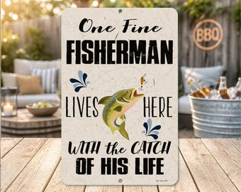 May include: A rectangular metal sign with the text "One Fine FISHERMAN LIVES HERE WITH the CATCH OF HIS LIFE." The sign features a jumping fish graphic and decorative blue accents. The background is a light beige color.