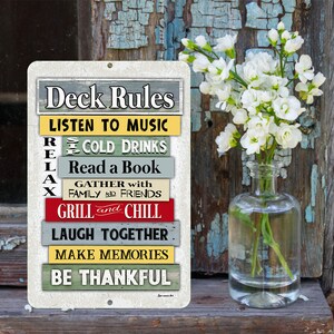 Może przedstawiać: Prostokątny metalowy znak z napisem "Deck Rules" na g&oacute;rze. Poniżej kolorowe poziome paski z frazami takimi jak "Listen to Music", "Cold Drinks" i "Grill and Chill". Przezroczysty szklany wazon zawiera białe kwiaty.