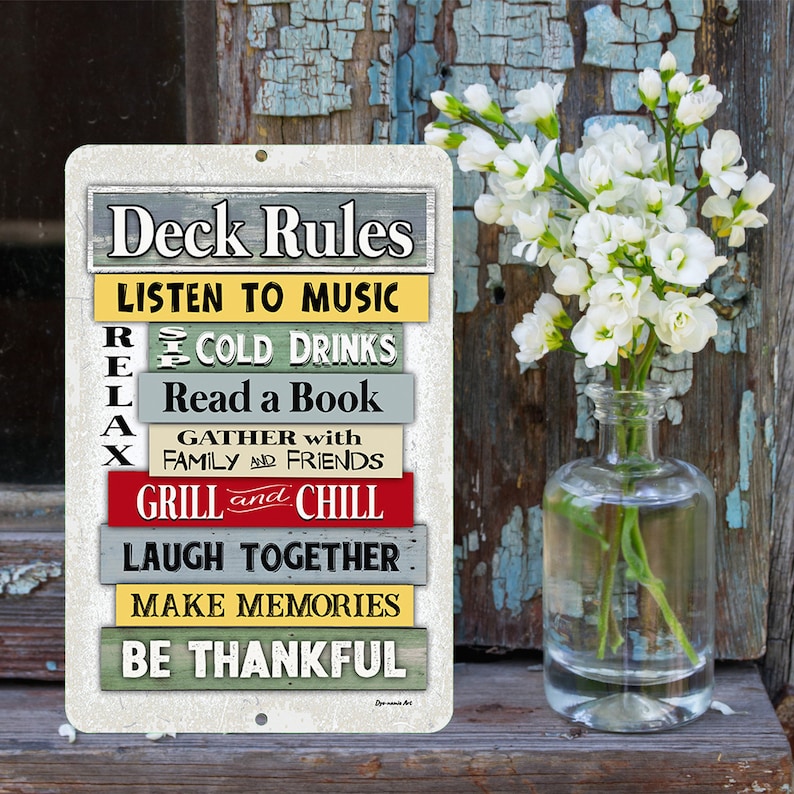 Może przedstawiać: Prostokątny metalowy znak z napisem "Deck Rules" na g&oacute;rze. Poniżej kolorowe poziome paski z frazami takimi jak "Listen to Music", "Cold Drinks" i "Grill and Chill". Przezroczysty szklany wazon zawiera białe kwiaty.