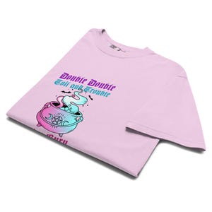 May include: A light pink t-shirt featuring a colorful graphic of a cauldron with smoke and the text "Double Double Toil and Trouble". The cauldron is in shades of pink, blue, and purple. The shirt is folded.