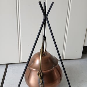May include: A copper pot with a rounded body and a lid, featuring a brass handle attached to the sides. The pot hangs from a black metal tripod stand constructed of three thin, intersecting rods that meet at the top to support the vessel.