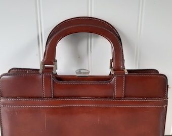 May include: A structured brown leather handbag featuring a semi-circular top handle, a rectangular body with visible stitching along the edges, and a silver-toned metal clasp closure at the center top. The bag has a smooth finish and clean lines.