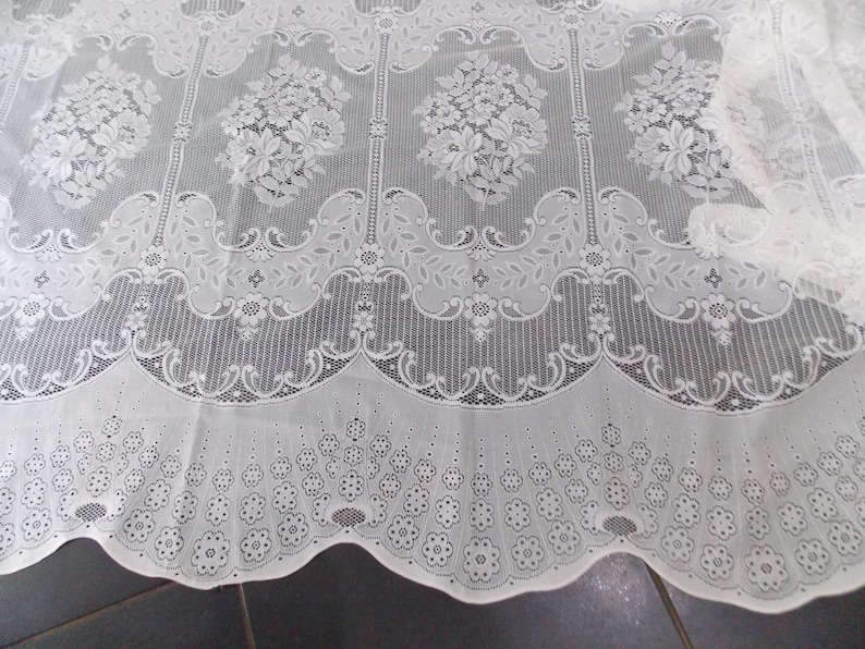 May include: White lace curtain panel with a delicate floral pattern. The lace is woven in a repeating pattern of small flowers and leaves. The bottom edge of the curtain is scalloped.
