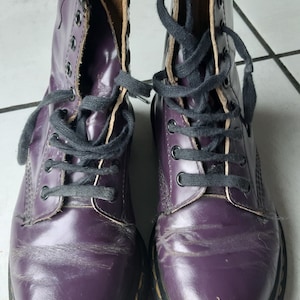 Puede incluir: Un par de botas Dr. Martens de cuero morado con cordones negros. Las botas tienen un detalle de costura amarilla alrededor de la suela.