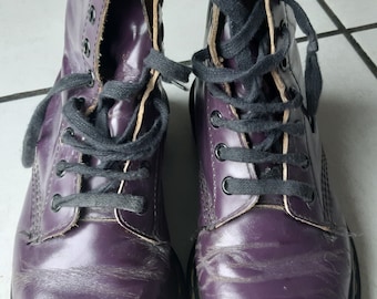 Puede incluir: Un par de botas Dr. Martens de cuero morado con cordones negros. Las botas tienen un detalle de costura amarilla alrededor de la suela.