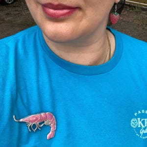 Op de afbeelding: Een felblauw t-shirt met een roze garnalenbroche op de borst, gecombineerd met een kleine aardbeivormige oorbel. Het t-shirt heeft witte tekst aan de rechteronderkant en de oorbel is een textuurrijke rode vrucht met een groen kroontje.