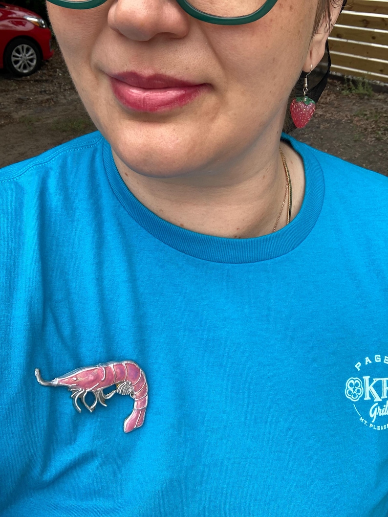 Op de afbeelding: Een felblauw t-shirt met een roze garnalenbroche op de borst, gecombineerd met een kleine aardbeivormige oorbel. Het t-shirt heeft witte tekst aan de rechteronderkant en de oorbel is een textuurrijke rode vrucht met een groen kroontje.