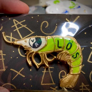 May include: A shrimp-shaped pin featuring a bright neon green body with gold outlines. The head displays a black and white yin-yang symbol with the letters V and B, while the tail section features the black letters L, O, and S.