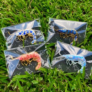 Op de afbeelding: Vier garnaalvormige emaille pins op zwarte achtergrondkaarten. E&eacute;n pin heeft blauwe bloemmotieven, &eacute;&eacute;n is donkerblauw, &eacute;&eacute;n is roze met gouden stippen en &eacute;&eacute;n is iriserend lichtblauw. Elke pin is afzonderlijk verpakt in doorzichtig plastic.