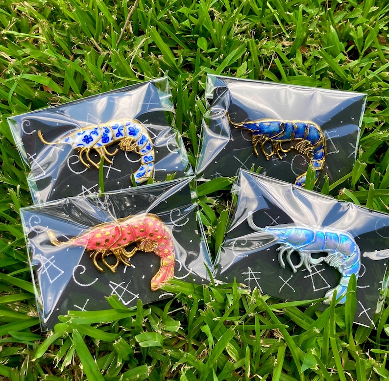 Op de afbeelding: Vier garnaalvormige emaille pins op zwarte achtergrondkaarten. E&eacute;n pin heeft blauwe bloemmotieven, &eacute;&eacute;n is donkerblauw, &eacute;&eacute;n is roze met gouden stippen en &eacute;&eacute;n is iriserend lichtblauw. Elke pin is afzonderlijk verpakt in doorzichtig plastic.