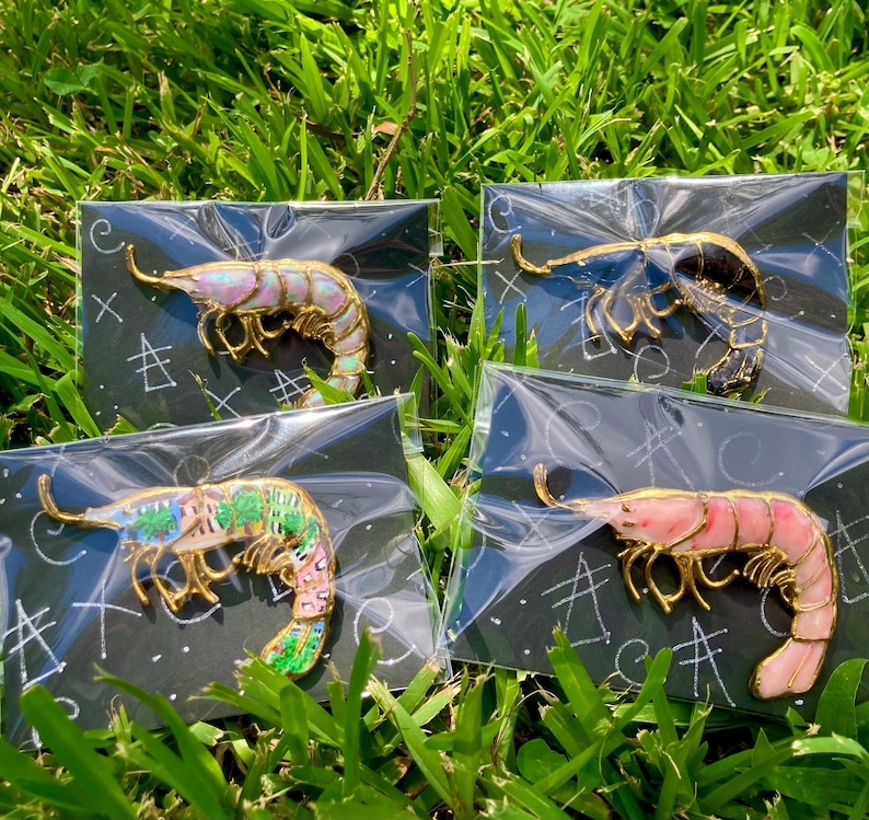 Op de afbeelding: Vier garnaalvormige emaille pins met goudkleurige metalen randen, elk individueel verpakt in doorzichtig plastic op zwarte achtergrondkaarten. De pins hebben verschillende kleurpatronen, waaronder iriserende, zwarte, groene en roze emaille ontwerpen.
