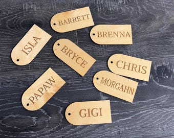 May include: Wooden name tags with rounded tops and a small hole. Each tag features a different name: Barrett, Brenna, Bryce, Chris, Gigi, Isla, Morgahn, and Papaw. The tags are light brown and sit on a dark wood surface.