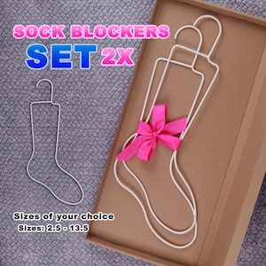 May include: Two white sock blockers in the shape of a sock, one with a pink bow. The sock blockers are in a cardboard box. The text "SOCK BLOCKERS SET 2X" is at the top of the image. The text "Sizes of your choice Sizes: 2.5 - 13.5" is at the bottom of the image.