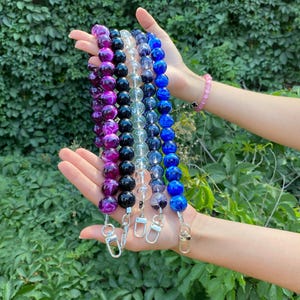 May include: A collection of beaded lanyards in various colors, including purple, black, clear, and blue. Each lanyard features large, round beads and a silver or gold-colored clasp. The lanyards are displayed against a backdrop of green foliage.