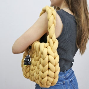 K&ouml;nnte beinhalten: Eine senfgelbe, grob gestrickte Tragetasche mit geflochtenen Henkeln. Die Tasche hat ein weiches, strukturiertes Aussehen und einen kleinen schwarz-wei&szlig;en Anh&auml;nger an der Seite. Die Tasche wird &uuml;ber der Schulter getragen.