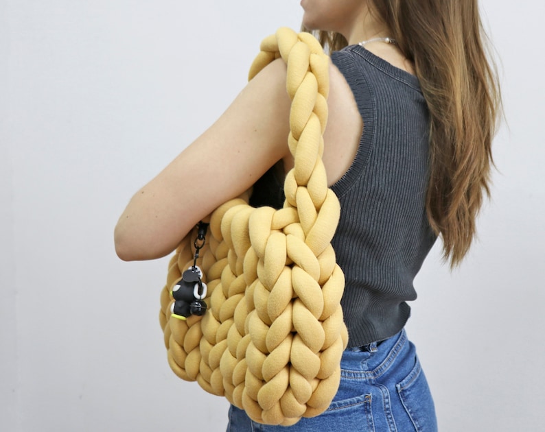 K&ouml;nnte beinhalten: Eine senfgelbe, grob gestrickte Tragetasche mit geflochtenen Henkeln. Die Tasche hat ein weiches, strukturiertes Aussehen und einen kleinen schwarz-wei&szlig;en Anh&auml;nger an der Seite. Die Tasche wird &uuml;ber der Schulter getragen.