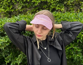 Pode incluir: Viseira de crochê rosa claro com aba curva. A viseira é usada por uma pessoa que usa uma camisa e jaqueta pretas. O fundo é uma parede de folhagem verde.