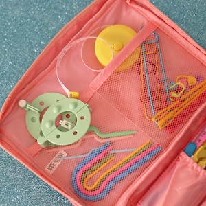May include: A pink crochet project bag with a mesh pocket containing colourful crochet hooks, a yellow tape measure, and a green yarn bowl. The bag has a zipper closure and the text "Made in China" printed on the side.