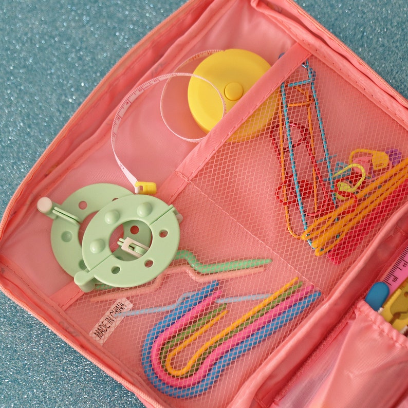 May include: A pink crochet project bag with a mesh pocket containing colourful crochet hooks, a yellow tape measure, and a green yarn bowl. The bag has a zipper closure and the text "Made in China" printed on the side.