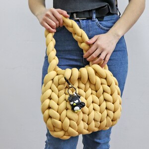 K&ouml;nnte beinhalten: Eine senfgelbe, grob gestrickte Handtasche mit geflochtenem Henkel. Die Tasche hat eine abgerundete Form und einen kleinen schwarz-wei&szlig;en Pinguin-Schl&uuml;sselanh&auml;nger an der Seite. Die Tasche wird von einer Person in Jeans gehalten.