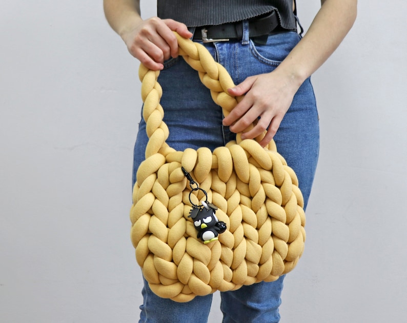 K&ouml;nnte beinhalten: Eine senfgelbe, grob gestrickte Handtasche mit geflochtenem Henkel. Die Tasche hat eine abgerundete Form und einen kleinen schwarz-wei&szlig;en Pinguin-Schl&uuml;sselanh&auml;nger an der Seite. Die Tasche wird von einer Person in Jeans gehalten.