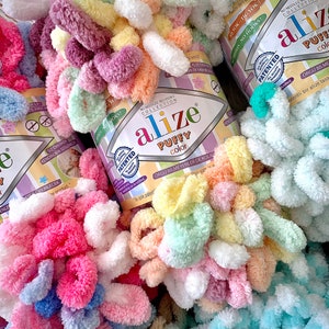 May include: Close-up of colorful Alize Puffy Color yarn skeins. The yarn is a soft, looped texture in shades of pink, yellow, green, purple, and white. The packaging features the brand name and product details.