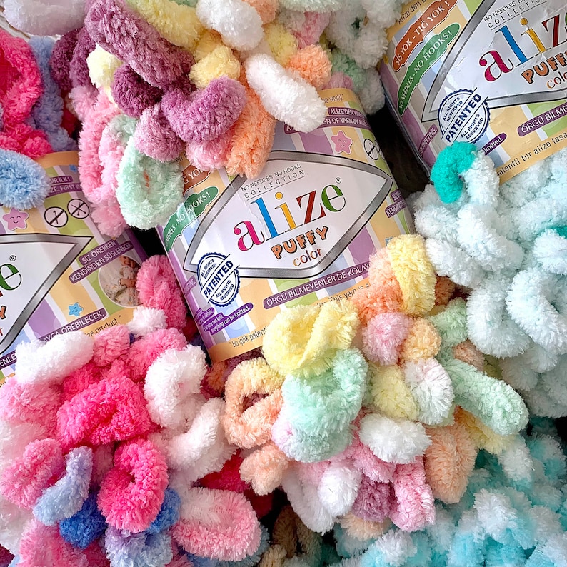May include: Close-up of colorful Alize Puffy Color yarn skeins. The yarn is a soft, looped texture in shades of pink, yellow, green, purple, and white. The packaging features the brand name and product details.