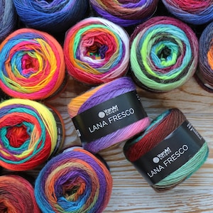 May include: A collection of round yarn balls in various vibrant, multi-coloured gradients. Two of the yarn balls feature black paper labels with the white text YarnArt UNIQUE LANA FRESCO printed on them, indicating the brand and product name.