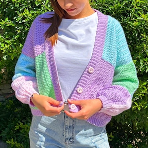 May include: A pastel-coloured crochet cardigan with a patchwork design. The open-front sweater features shades of purple, blue, green, and pink, with long sleeves and three white buttons. A unique, handmade garment.