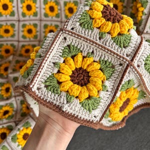 Puede incluir: Una manta de ganchillo hecha a mano con un patr&oacute;n repetido de girasoles. La manta est&aacute; hecha de hilo crema, amarillo, marr&oacute;n y verde. Los girasoles son el punto focal, con p&eacute;talos y centros detallados. La manta es cuadrada y parece suave y acogedora.