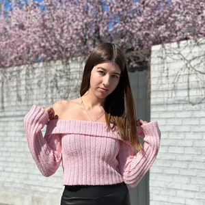 Puede incluir: Un suéter rosa de hombros descubiertos con mangas largas y textura de punto de canalé.