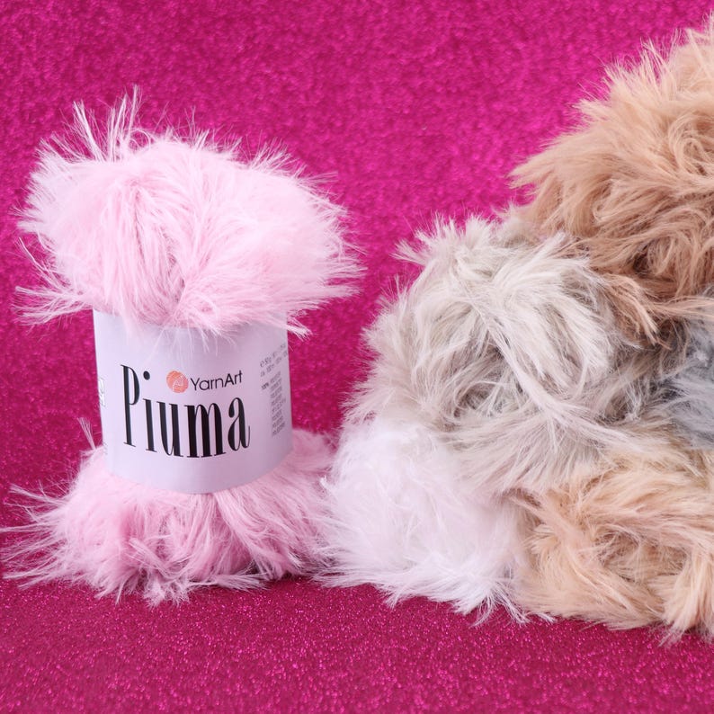 May include: A close-up of pink, white, and tan fluffy yarn. A pink skein of YarnArt Piuma yarn is in the foreground. The background is a bright pink, glittery surface. The yarn appears soft and textured, suitable for knitting or crochet projects.