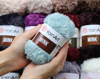 May include: A skein of light blue faux fur yarn with a white and brown paper label reading YarnArt MINK and www.yarnart.info. The yarn has a soft, fuzzy texture and is surrounded by other skeins of yarn in various colors including white, purple, and dark red.
