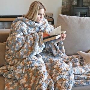 May include: A person is sitting on a sofa wearing a large, soft, blue, white and beige knitted blanket with sleeves. The blanket is made of chunky yarn and has a loose, drapey texture. The person is reading a book.