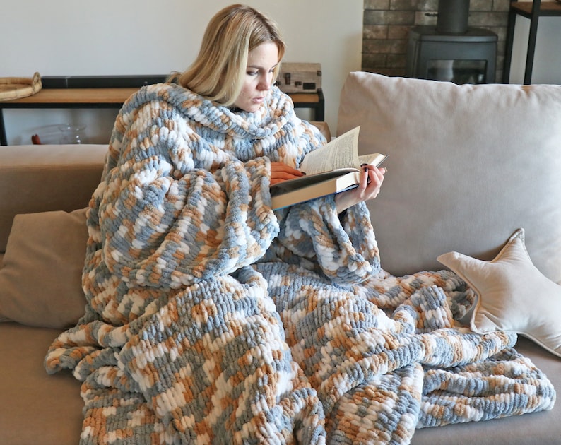 May include: A person is sitting on a sofa wearing a large, soft, blue, white and beige knitted blanket with sleeves. The blanket is made of chunky yarn and has a loose, drapey texture. The person is reading a book.