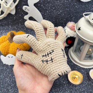 May include: A beige crochet hand with black stitched details, perfect for Halloween decor. The hand features detailed stitching and is designed to resemble a zombie hand. Other Halloween decorations are in the background.